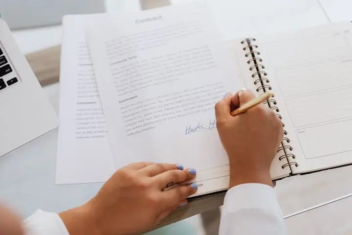 A person signs a contract with a pen next to an open planner on a desk, with other documents and a laptop partially visible.