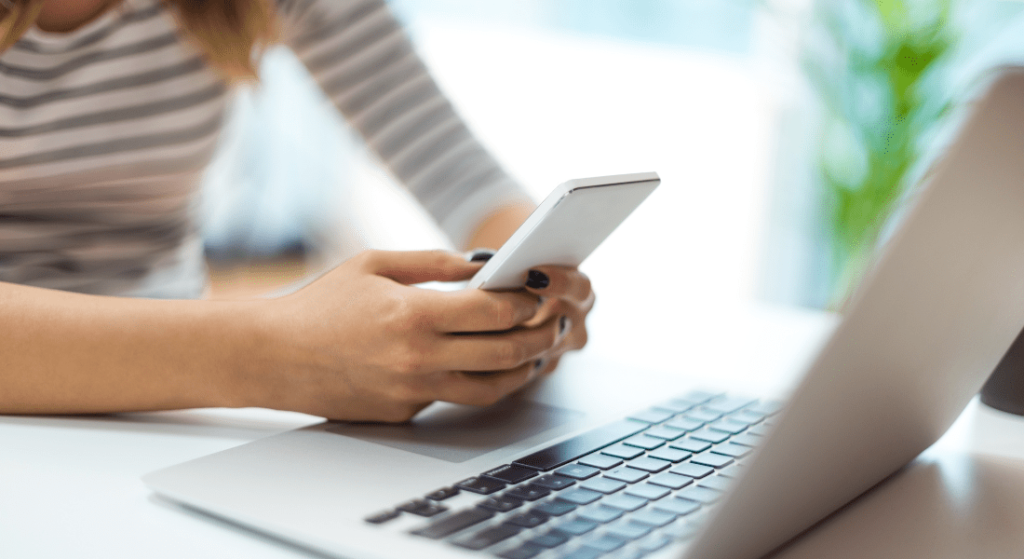 A person is using a smartphone while sitting at a desk with an open laptop.