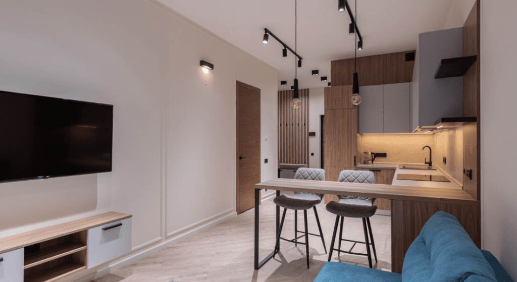 Modern apartment interior with a wall-mounted TV, a kitchen area featuring wood cabinets, a small bar with two stools, and a blue sofa. Track lighting on the ceiling. Average Rent in Sango
