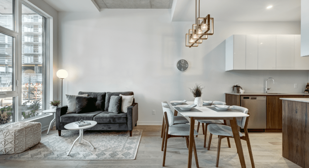 Modern open-plan living and dining area with grey sofa, white dining table with four chairs, pendant lights, and large window. Neutral tones and minimalistic decor. Average Rent in Greenwood, Clarksville, TN.