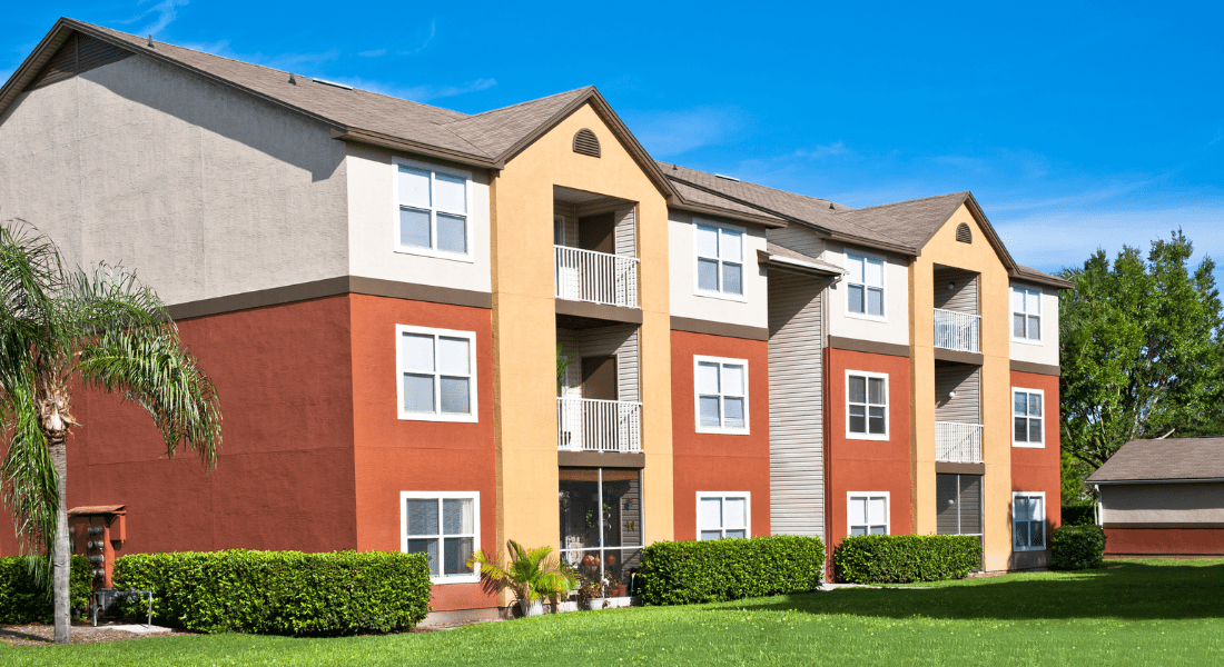 A three-story apartment building with red and beige exterior, balconies, and a manicured lawn under a bright blue sky. rental property greenwood clarksville tn.