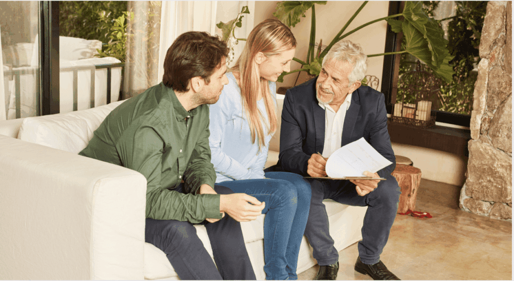 Three people sitting on a couch engaged in conversation; an older man is showing paperwork to the younger pair.
