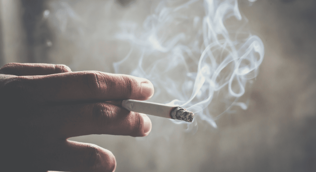 Hand holding a lit cigarette with smoke curling upwards against a blurred background. Can Tenant Smoke In Rented Property?