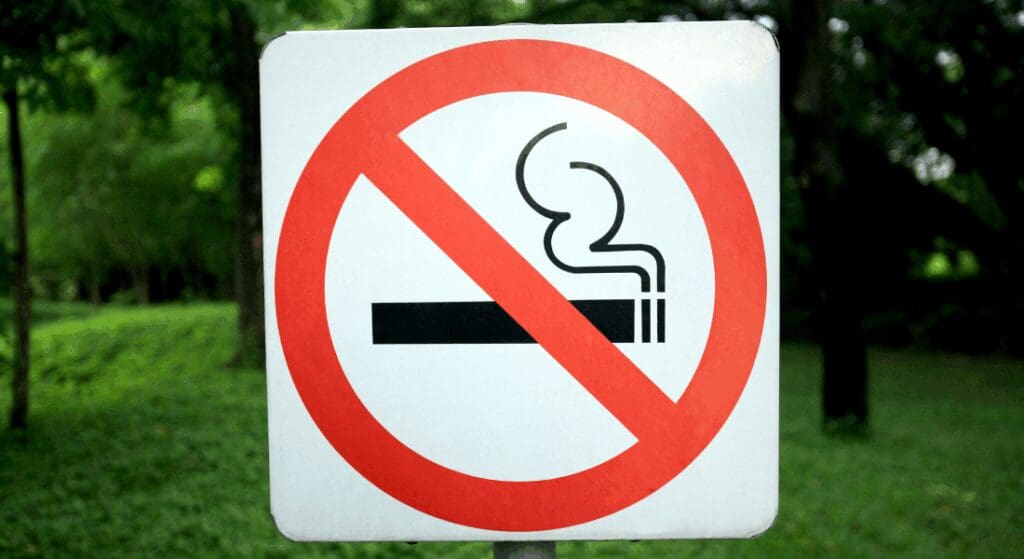 A no smoking sign with a cigarette graphic inside a red circle and diagonal line, posted outdoors with green trees and grass in the background.