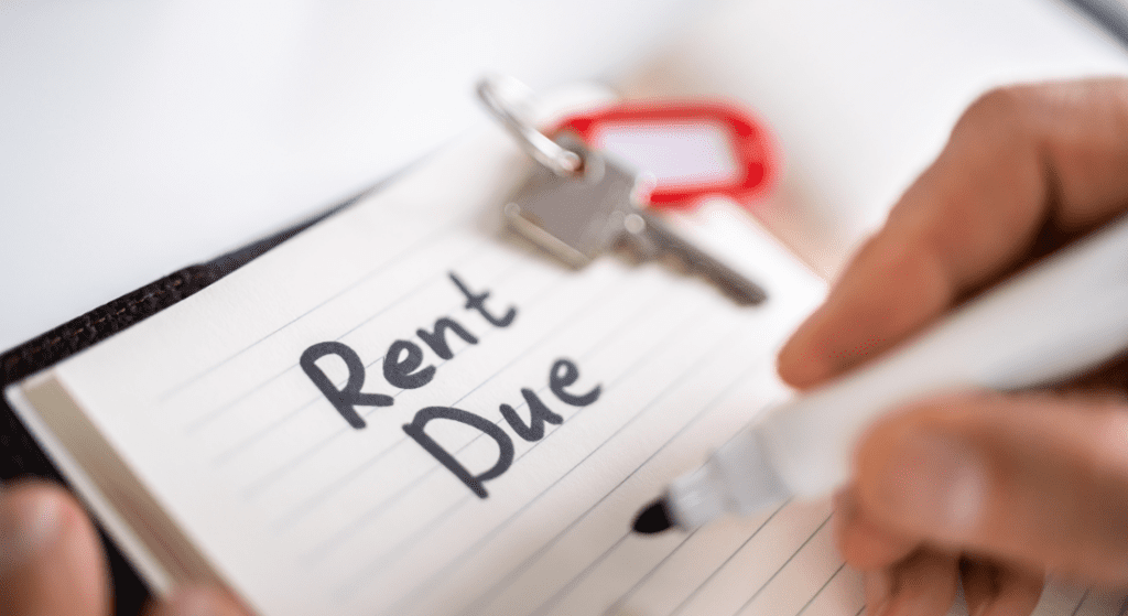 Close-up of a hand writing "Rent Due" on a notepad with a marker. A key with a red keychain is visible in the background.