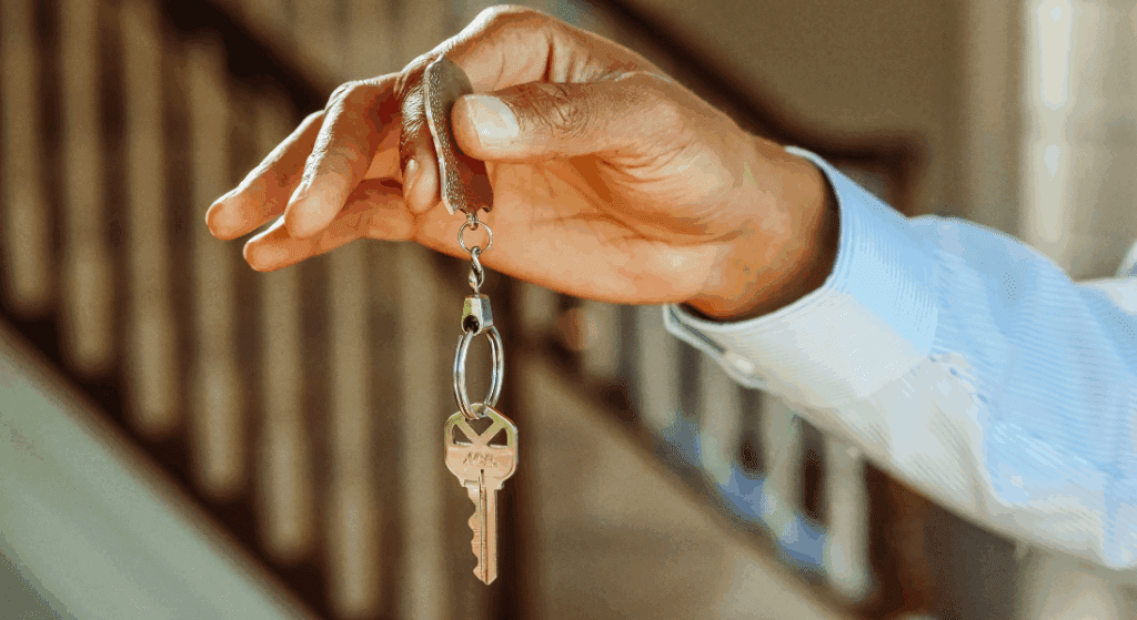 A person in a blue shirt holds a house key on a keychain inside a building, with a staircase visible in the background.
