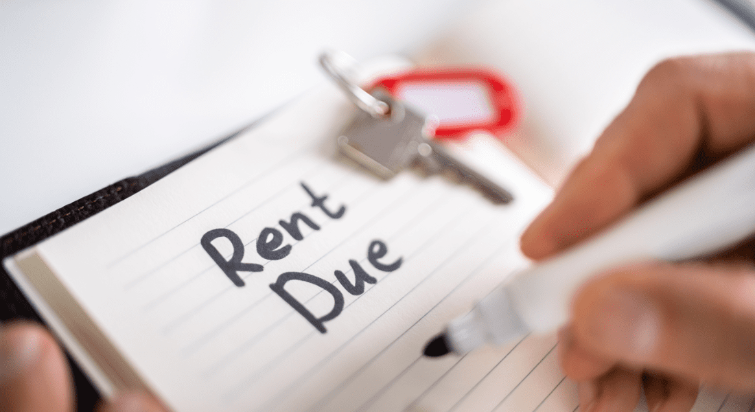 Close-up of a hand writing "Rent Due" on a notepad with a marker. A key with a red keychain is visible in the background.