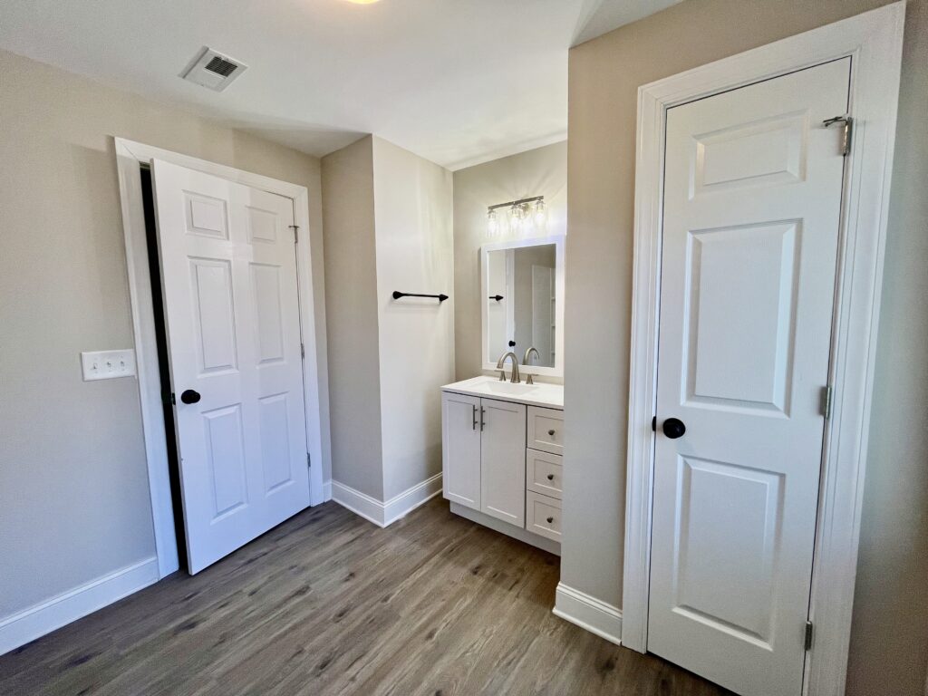 A modern bathroom with light gray walls, wooden flooring, a white vanity with double sinks, a mirror, and overhead lighting. Two white doors are visible, one open and one closed.