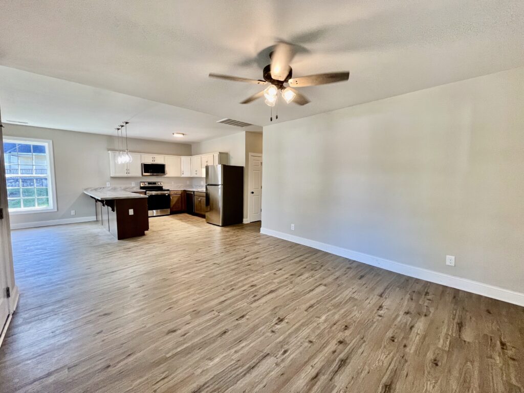 Modern open-concept kitchen and living room with stainless steel appliances, white cabinets, wood-look vinyl flooring, and ceiling fan lighting.
