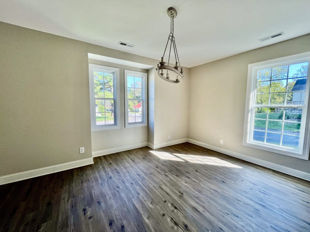 Bright dining room with large windows, modern chandelier, and dark wood-look vinyl flooring in a neutral-colored interior.