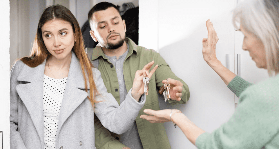 A young couple hands over keys to an older woman at a doorway, with the woman holding up her hand in a dismissive gesture.