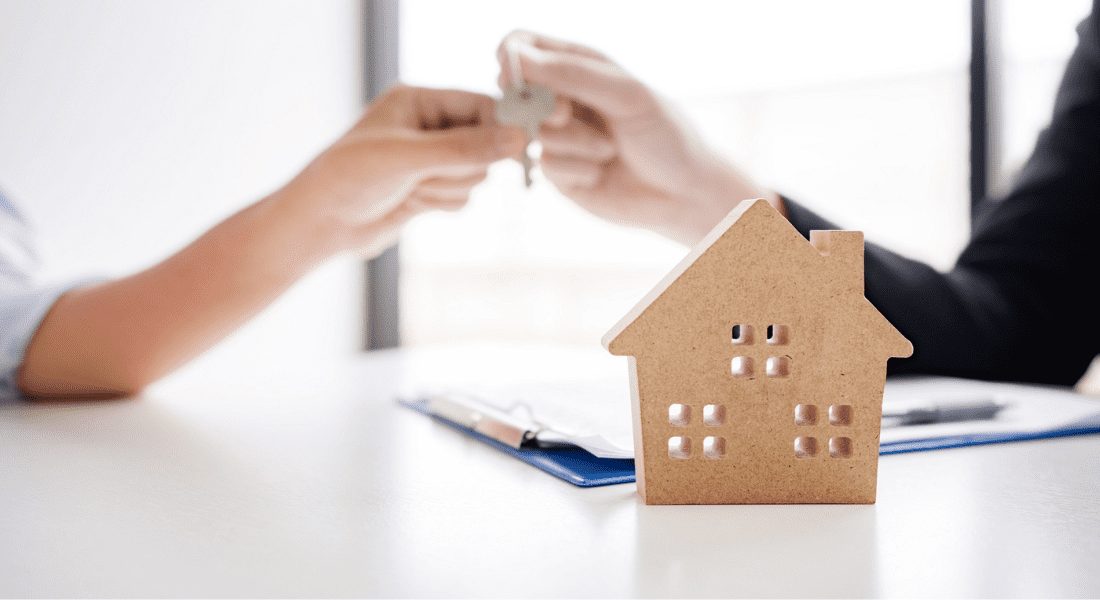 A small model house on a table, with two people exchanging keys in the background and a clipboard with documents nearby.