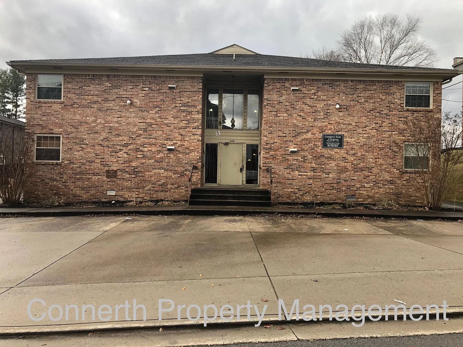 Two-story brick apartment building with a central glass entryway, concrete steps, and bare trees in front. "Connerth Property Management" text overlaid at the bottom.