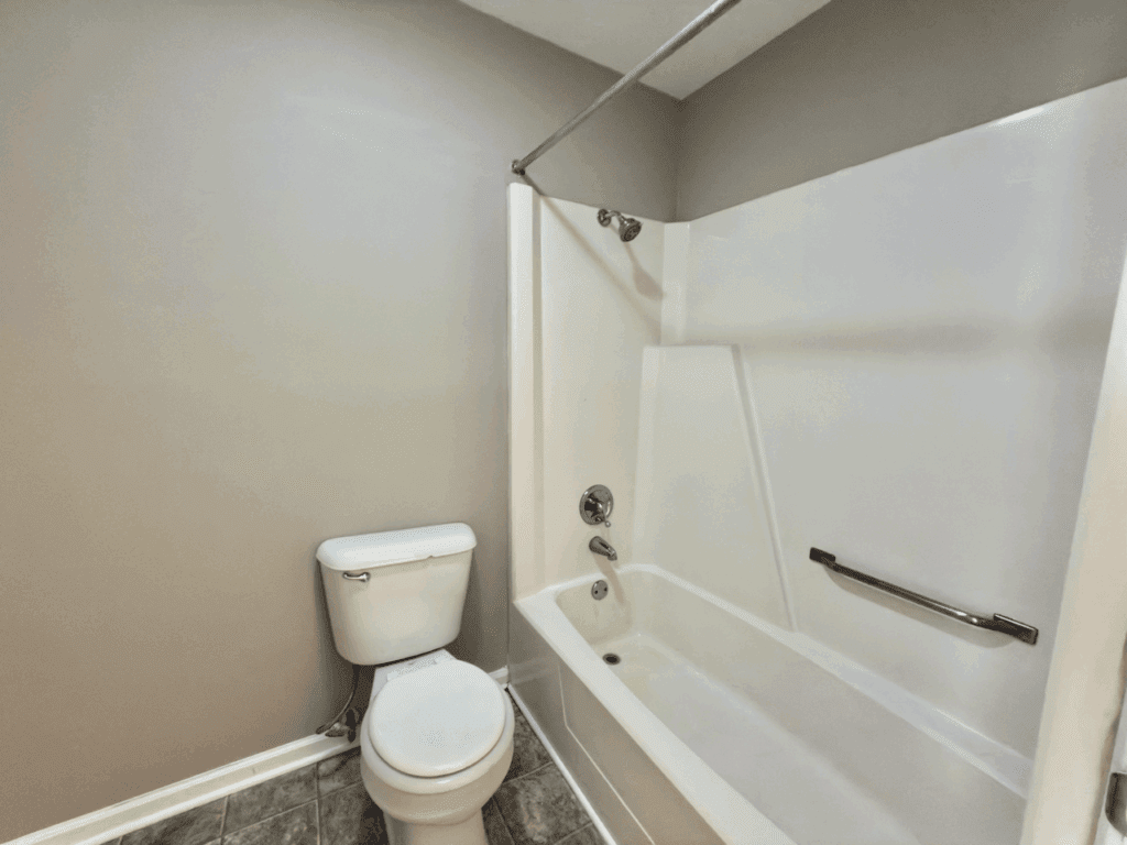 Bathroom with a white toilet and a white bathtub-shower combination, featuring a grab bar and beige walls.