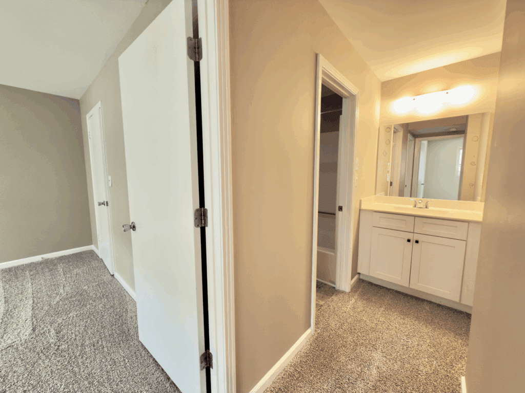 View of a hallway with a carpeted bedroom on the left, an open door, and a bathroom with a vanity and mirror on the right. Walls are painted beige and lighting is warm.