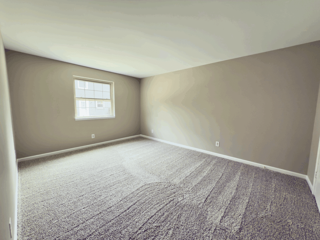 An empty room with beige walls, a single window letting in daylight, and freshly vacuumed light brown carpet.