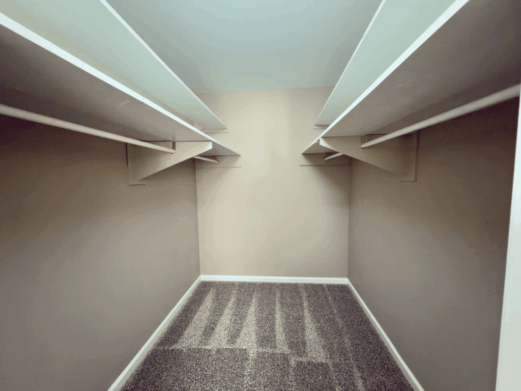 Empty walk-in closet with beige walls, carpeted floor, and two rows of white shelves and hanging rods on both sides.