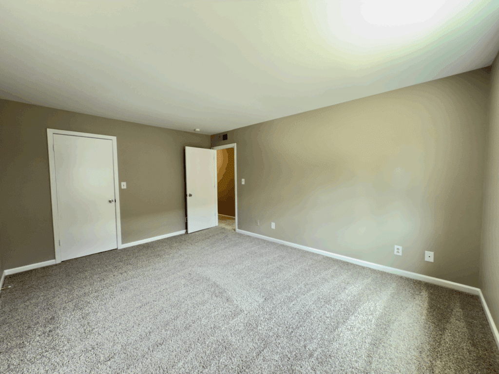 Empty carpeted room with beige walls, two closed white doors, one open door, white trim, and an overhead light.