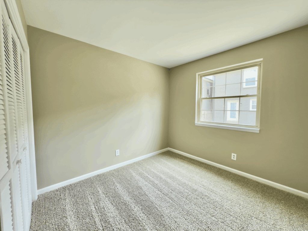 Empty room with beige walls, carpeted floor, a window letting in natural light, and a closed closet with white louvered doors.