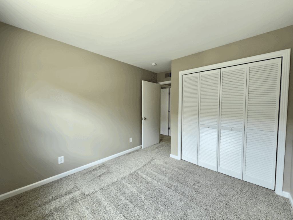 Empty bedroom with beige walls, carpeted floor, an open white door, and a closed white closet with louvered doors.