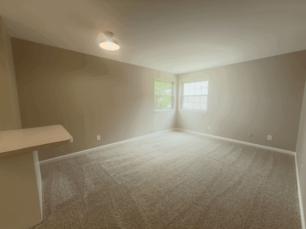 Empty carpeted room with beige walls, two windows letting in natural light, and a ceiling light fixture. A countertop is visible on the left side of the image.