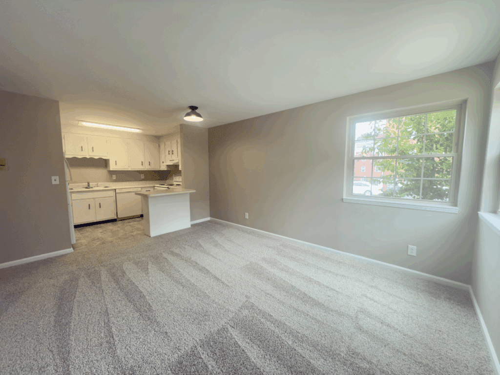 Unfurnished living area with carpeted floor, neutral walls, a large window, and an open view of a small kitchen with white cabinets.