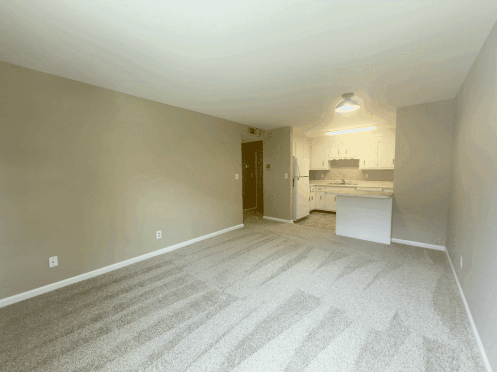 Unfurnished living space with carpeted floor, beige walls, and an open view of a white kitchen area with cabinets, refrigerator, and overhead light.