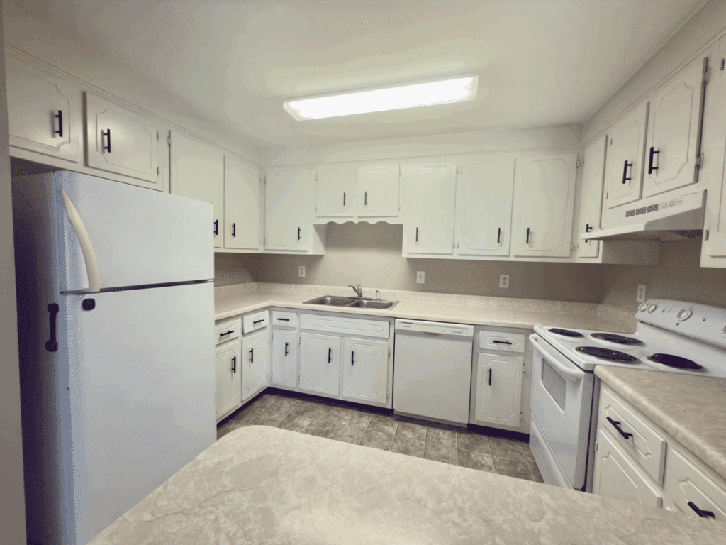 White kitchen with upper and lower cabinets, a refrigerator, stove, dishwasher, double sink, and beige countertops under fluorescent lighting.