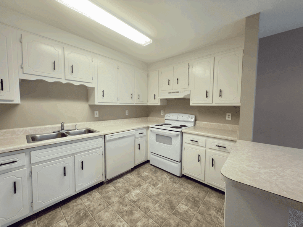 Kitchen with white cabinets, electric stove, dishwasher, double sink, and beige countertops. Overhead fluorescent light and neutral tile flooring.