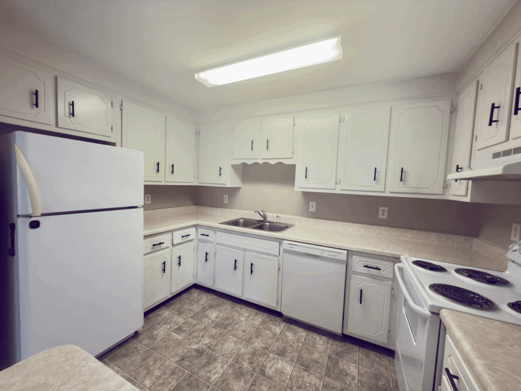 Small kitchen with white cabinets, a white refrigerator, stove, dishwasher, double sink, and fluorescent ceiling light. The floor has patterned tiles and counters are beige.