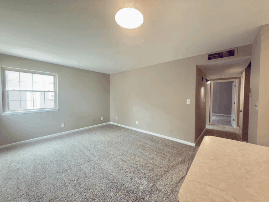 Empty carpeted room with beige walls, a window on the left, overhead light, air vent on the ceiling, and a hallway leading to another area.