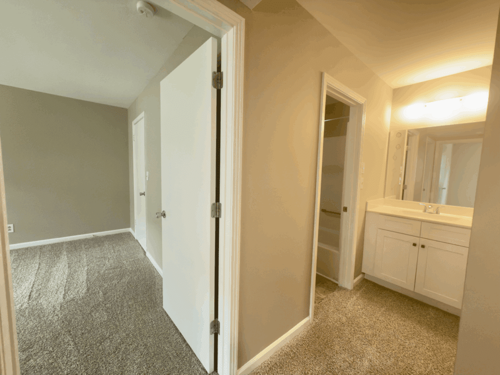 View of a beige carpeted hallway leading to a bedroom on the left and a white bathroom vanity with a mirror and lights on the right.