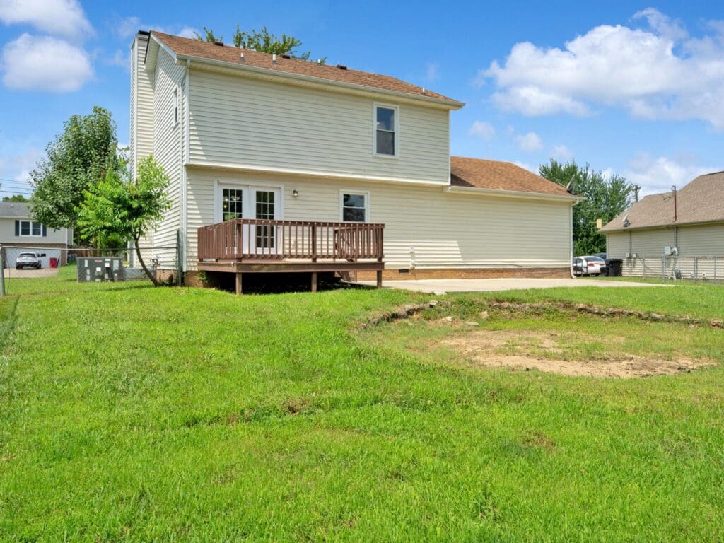 Two-story beige house with a small raised wooden deck overlooks a grassy backyard with a patch of bare dirt under a blue sky with scattered clouds.