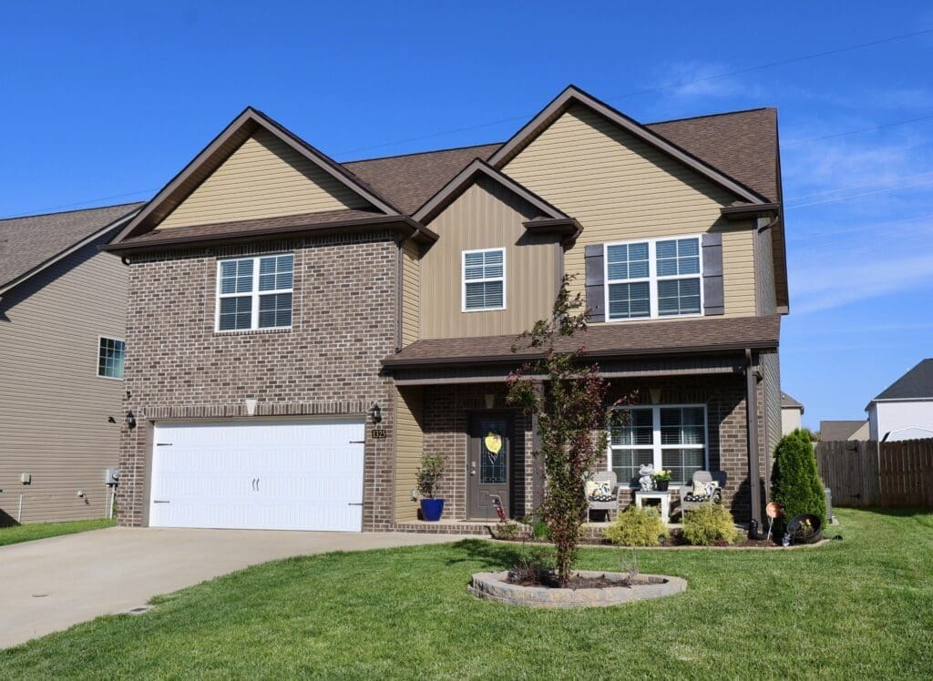 Two-story suburban house with brick and tan siding, white garage door, small front porch with chairs, and a landscaped front yard on a sunny day.