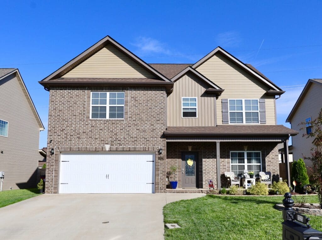Two-story suburban house with brick and tan siding exterior, white garage door, and a small front porch with chairs, set on a concrete driveway and green lawn.