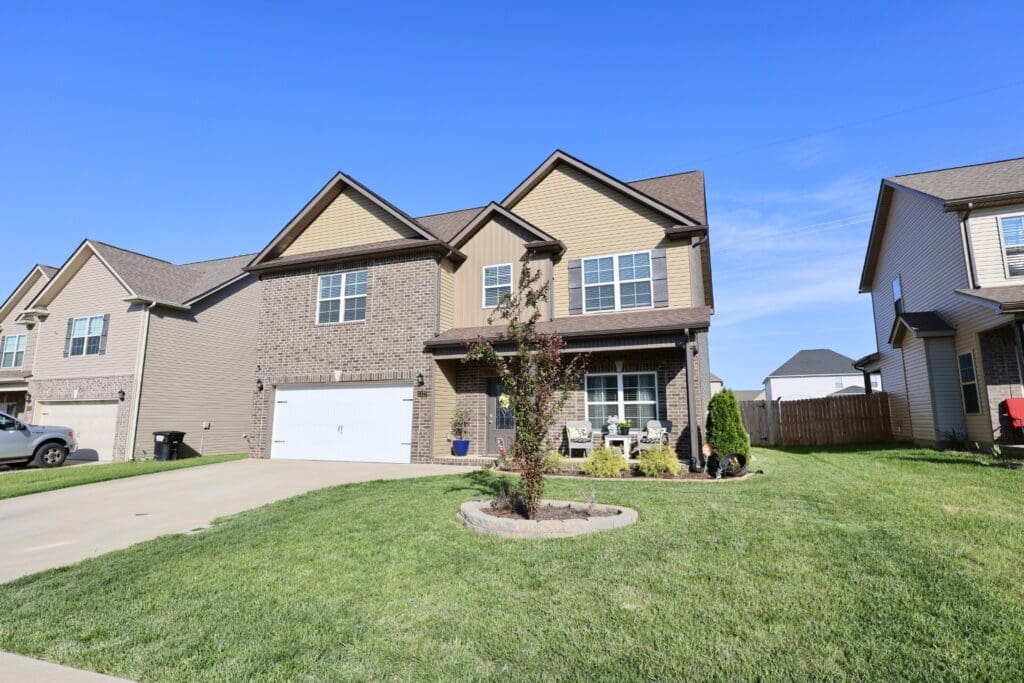 Two-story suburban house with a brick and siding facade, double garage, manicured lawn, and small front garden under a clear blue sky.