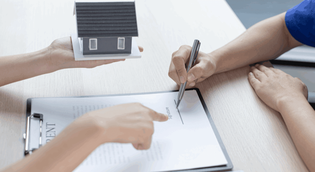 A person holds a model house while another person signs a document on a clipboard, with a third hand pointing at the signature line.