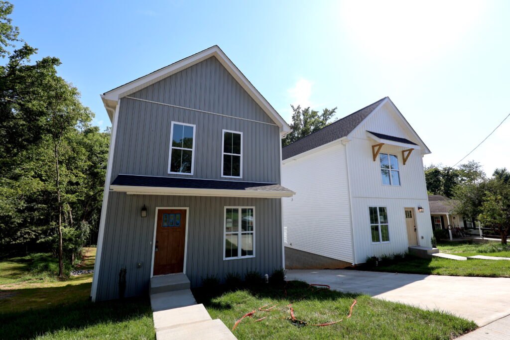 Two modern, two-story houses with pitched roofs sit side by side, one gray and one white, with concrete driveways and green lawns in a residential neighborhood.