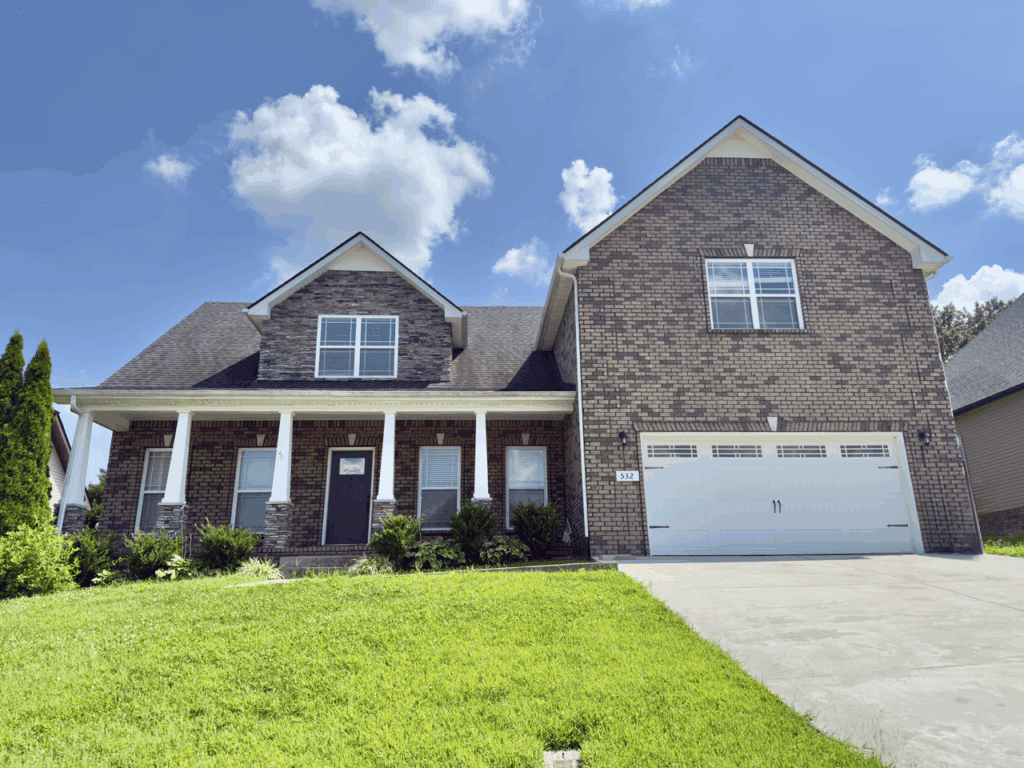 A two-story brick house with white trim, a covered front porch, and a double garage, situated on a lawn under a partly cloudy sky.