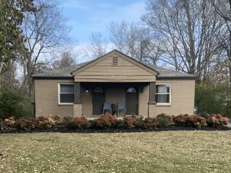 A small, single-story house with two front doors, a covered porch, and a row of shrubs and flowers in the front yard. Leafless trees are visible in the background.