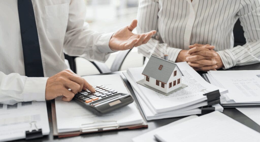 Two people in business attire discuss documents with a calculator and a small model house on the table, possibly reviewing real estate or mortgage information.
