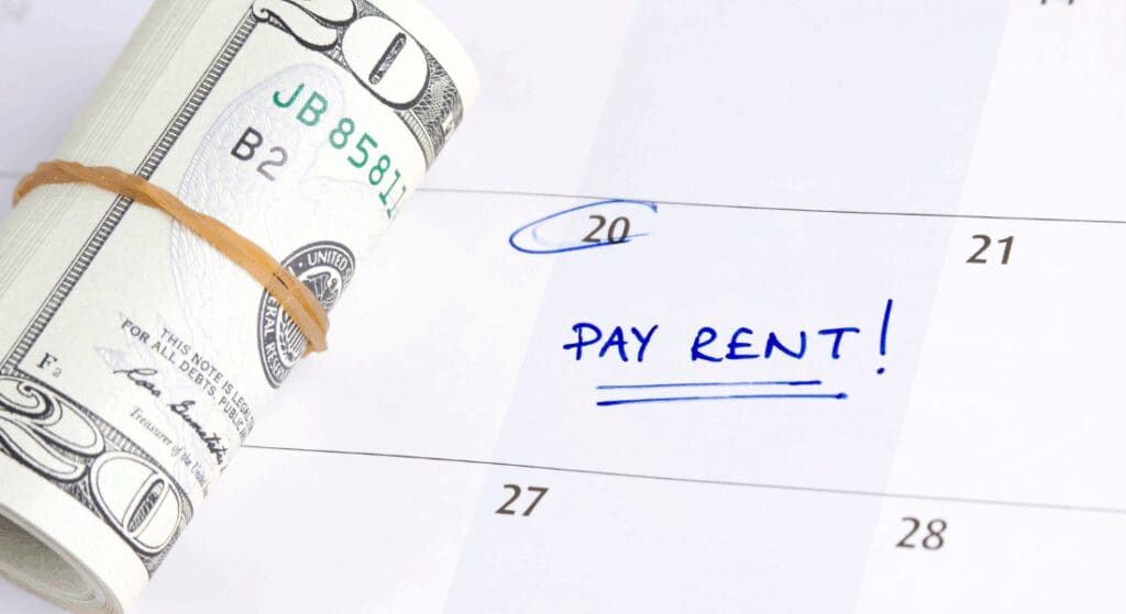 A rolled-up $100 bill next to a calendar with the date circled and the words "PAY RENT!" written in blue ink.