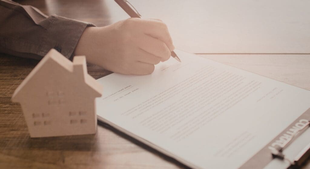 A person signs a contract on a clipboard next to a small wooden house model on a wooden table.