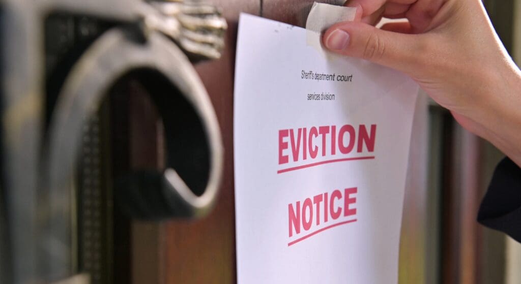 A hand tapes an eviction notice to a door. The notice has the words "EVICTION NOTICE" printed in bold red letters.