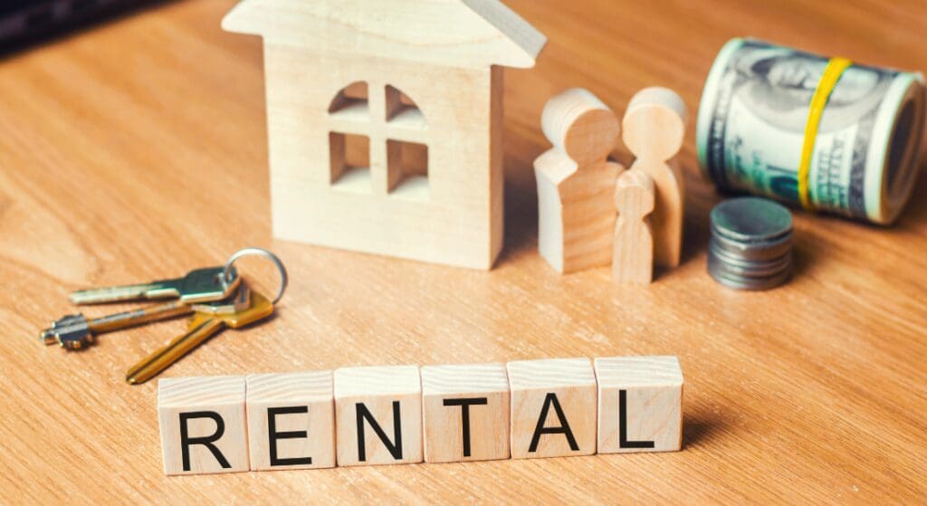 Wooden blocks spell "RENTAL" on a table with a house model, family figures, keys, coins, and a roll of dollar bills, symbolizing property rental or real estate investment.