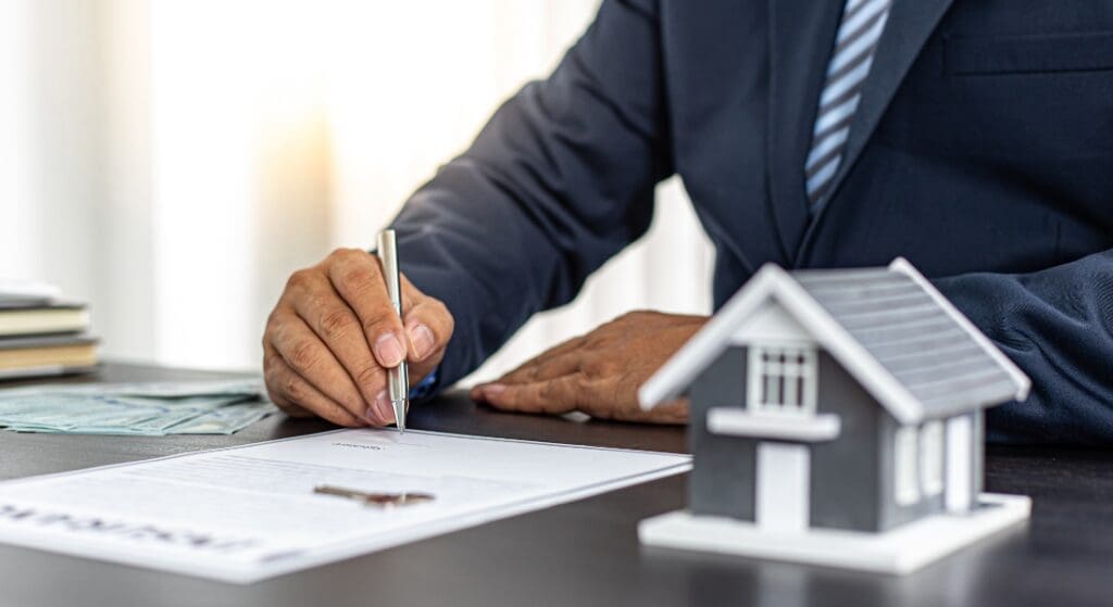 A person in a suit signs a document at a desk, with a model house, keys, and a stack of cash visible nearby.