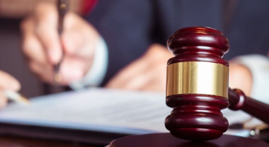 A wooden judge's gavel rests on a sound block, with a person writing on a document in the blurred background.
