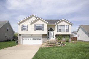 A two-story beige house with white trim, a double garage, front steps, manicured lawn, and a small garden in the front yard.