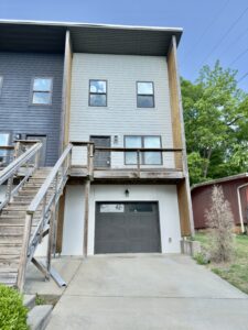 Three-story townhouse with gray siding, two upper windows, a small balcony with wood accents, a single garage door, and a concrete driveway. Trees and a red shed are in the background.