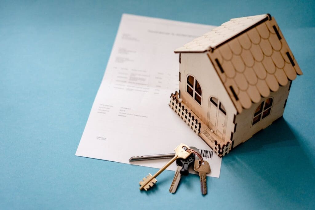 A small wooden model house, a set of keys, and a document are arranged on a light blue surface.